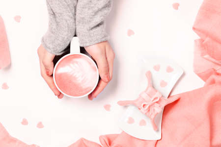 Female hands in grey  knit holding cup of cappuccino. Gift alike dessert with heart shape sprinkles. Flat lay. Valentines concept. Pink textile. Horizontal. Living coral theme - color of the year 2019の写真素材