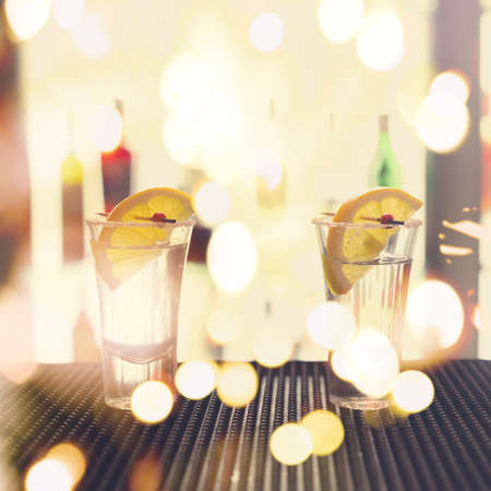 Two tequila shots with lemon on a bar ribber mat against highlighted glass wall. Shallow DOF, unusual light and toned with festive bokeh lightsの写真素材