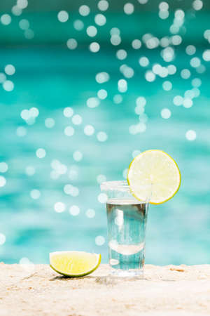One tequila shots with sliced lime on the beach. Transparent alcohol. Hanging out. Vertical with gentle festive holiday bokehの写真素材