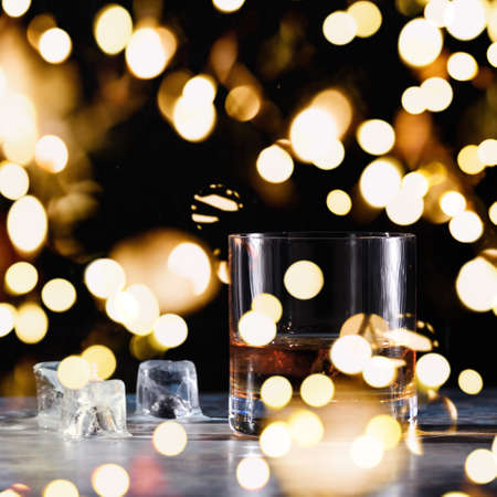 Glass of whiskey on the rocks with some ice on the table. Concept of hard liquor. Square with festive holiday bokehの写真素材