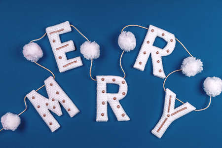 White and golden knitted garland with word MERRY and white pompons on classic blue. Holiday celebration concept, Christmas cardの写真素材