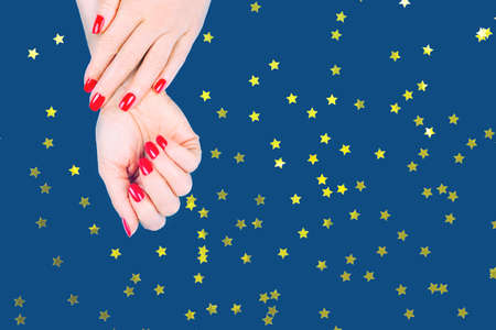 Woman hands with stylish red manicure on blue background. Holiday, party and beauty concept. Horizontal with festive golden star shaped confettiの写真素材