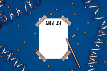 Isolated white card with Guest List wording and golden pen with festive golden ribbons and confetti. Flat lay background in gold and classic blue colors for any celebration and party occasionの写真素材
