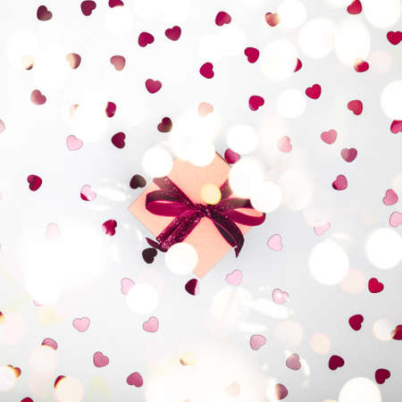 Craft box with dark red velvet ribbon bow and glitter heart confetti. Valentine day and eco-friendly wrapping concept. Trendy minimalistic flat lay design background. Square with festive bokeh lightsの写真素材