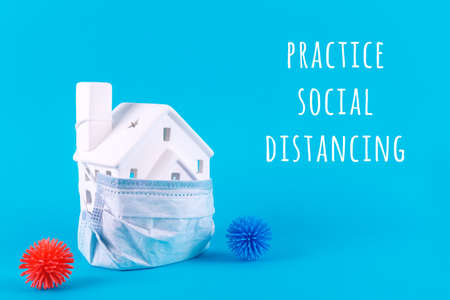 Cute little house covered with medicine mask and plastic balls viruses on the blue with Practice Social Distancing wording. Epidemic, social isolation, coronavirus COVID-19 concept. Option with textの写真素材