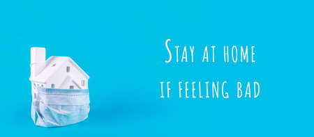 Cute little house covered with medicine mask on the blue background with Stay at home if feeling bad wording. Epidemic, social isolation, coronavirus COVID-19 concept. Option with textの写真素材
