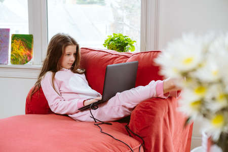Preteen girl enjoyes homeschooling in comfy pijama and cozy sofa chair. Remote education, virus protection, social distancing and lockdown concept. Flowers at the frontの写真素材