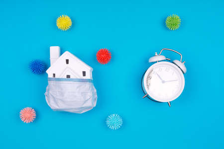 Cute little house covered with medicine mask, clock and plastic balls as viruses on the blue background. Epidemic, social isolation, coronavirus COVID-19 concept. Horizontalの写真素材