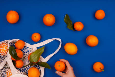 Woman taking mandarine from mesh cotton reusable bag on classic blue drop. Eco friendly shopping, ecology and healthy food concept, vitamin C citrus fruit. Place for text over fruit patternの写真素材