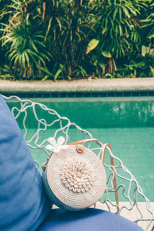 Trendy round crossbody straw bag with seashells and flower on the beautiful lounge chair near a pool. Summer outfit, vacation, holiday concept. Travel background with place for textの写真素材