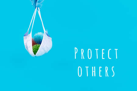 Hand in latex glove holds disposable mask with plastic balls as viruses on the blue background with Protect others wording. Epidemic, social isolation, coronavirus COVID-19 concept. Option with textの写真素材
