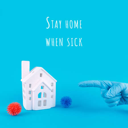 Hand in disposable glove points to cute little house with Stay home when sick wording. Epidemic, respoiratory desease, social isolation, coronavirus COVID-19 concept. Square format with place for textの写真素材