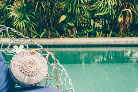 Trendy round crossbody straw bag with seashells and flower on the beautiful lounge chair near a pool. Summer outfit, vacation, holiday concept. Travel background with place for textの写真素材