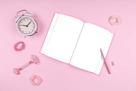 Journal with stylish pen, jade face roller, white clock and pink accessories. Beauty time, selfcare and beauty accessories concept. Place for textの写真素材