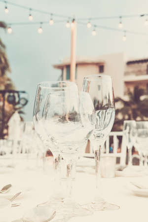 Festive serving with glasses for an outdoor celebration with garland lamps and palms at the background. Wedding, anniversary, birthday dinner and party. Toned image with place for textの写真素材