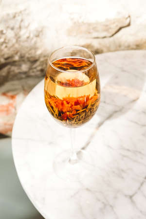 Blooming flower tea served in a wine glass, white marble table at the trendy cafe. Luxury tea party, creative tea serving concept. Place for textの写真素材