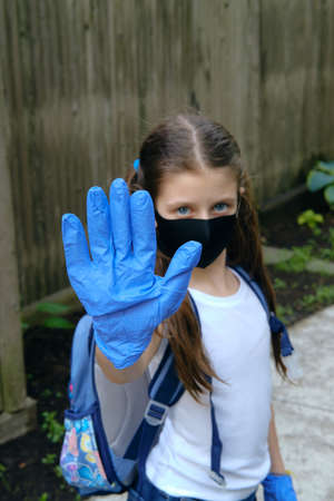 Elementary school student in a cloth dust mask and gloves with backpack in the street makes stop sign. Preteen girl is going to school in new normal. Education, coronavirus, back to school conceptの写真素材