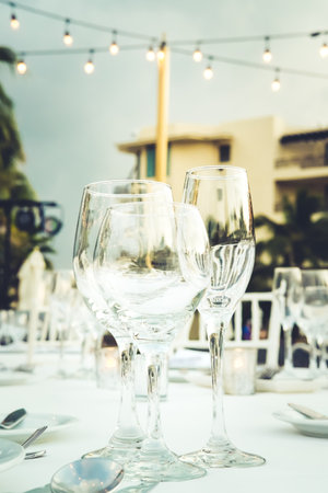 Festive serving with glasses for an outdoor celebration with garland lamps and palms at the background. Wedding, anniversary, birthday dinner and party. Toned image with place for textの写真素材