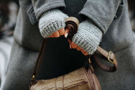 A girl in a coat and knitted gloves is holding a leather bag. Close-up of hands. A woman expects,...