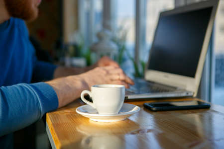 Close-up of mens hands, typing on a laptop, drinking coffee, next to the phone. A cup of coffee is empty. Close-up. Sunlightの写真素材