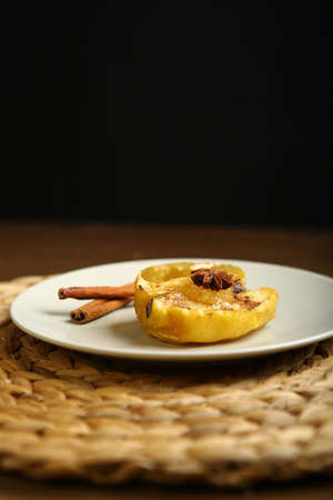 On the plate is a baked apple, near cinnamon, anise. Wicker substrate and wooden table, composition on a black background.の写真素材