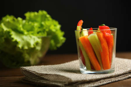 Pepper, carrots, celery, raw cucumber are cut into strips. In a glass beaker. Next to it are lettuce leaves, basil. On a background of cloth, burlap, wooden table, black background.の写真素材