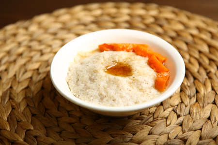 Low-calorie porridge for a healthy diet and lifestyle. Barley with honey and bits of orange pumpkin in a deep white plate. Dark background, wooden table and wicker standの写真素材