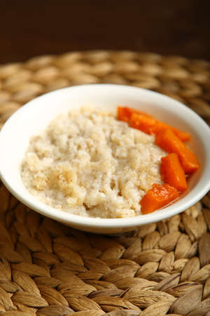 Low-calorie porridge for a healthy diet and lifestyle. Barley with honey and bits of orange pumpkin in a deep white plate. Dark background, wooden table and wicker standの写真素材