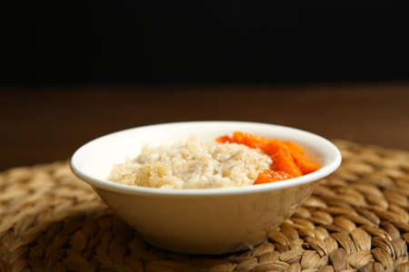 Low-calorie porridge for a healthy diet and lifestyle. Barley with honey and bits of orange pumpkin in a deep white plate. Dark background, wooden table and wicker standの写真素材