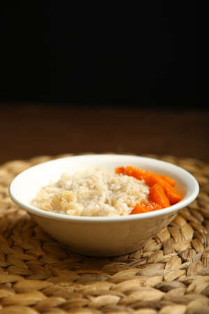 Low-calorie porridge for a healthy diet and lifestyle. Barley with honey and bits of orange pumpkin in a deep white plate. Dark background, wooden table and wicker standの写真素材