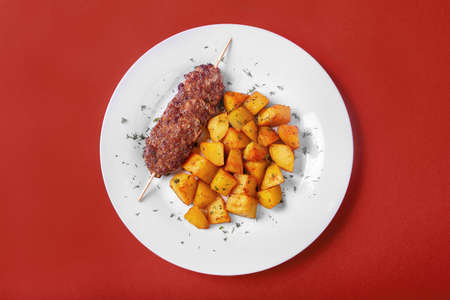 Meat kebab from pork, beef with fried potatoes on a white plate. Red monophonic background. A hearty meal, dinner. Dish for the menu. View from aboveの写真素材
