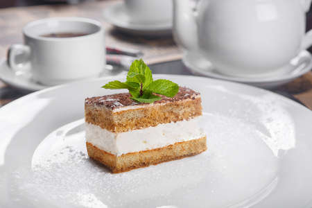 Rectangular piece of Tiramisu with a sprig of mint on a white plate. In a low key, in the background a cup of tea and a kettle, cutlery. Serving, serving dishes in a restaurant, cafe, barの写真素材