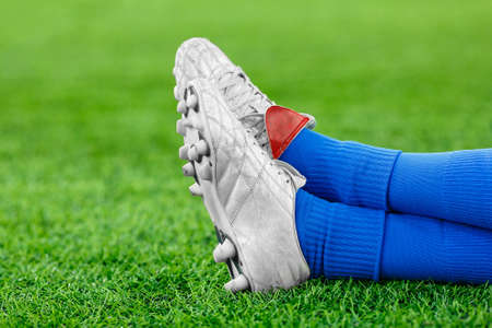 Legs of a player in football, an athlete in blue gaiters and white studded sneakers on a green lawn. Place for the inscription, rest, break, timeout, fatigue, sits, lies. Close upの写真素材