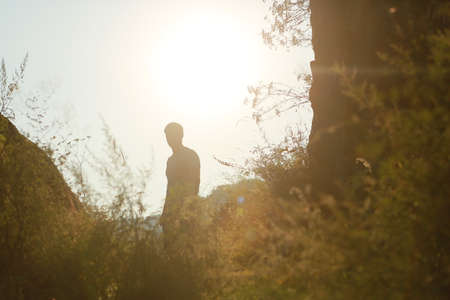 Silhouette of a man in a hollow, among mountains, grass, in a valley, sunlight behind, sunset, hiking alone. A man walks, leads a healthy lifestyle, stopped, standsの写真素材