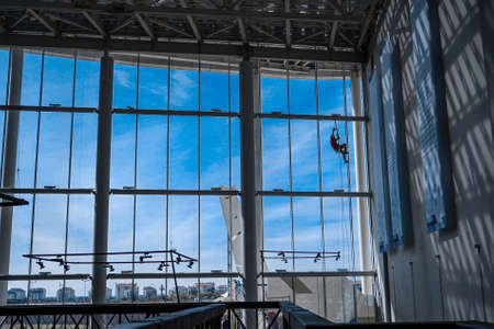 Industrial climber hangs from ropes inside shopping center or exhibition hall building against the blue sky, plenty of free space to signの写真素材