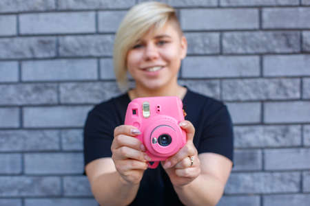 stylish large girl in a black t-shirt smiles and takes pictures on a pink camera against the wall