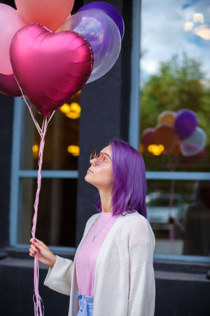 Young lady with violet hair in pink glasses, white blouse and pink t-short looking up at buch of...
