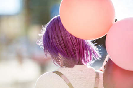 Woman with violet hair in white cardigan with bunch of colored balloons standing turn her back outdoor in cityの写真素材