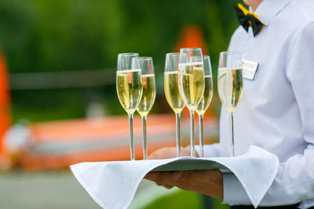 A waiter in white shirt is serving a few glass of champagne on the tray with white napkin
