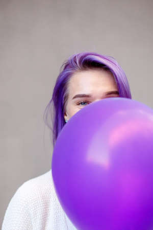 Female with violet hair in white blouse cover her face with one purple airballoon standing in front gray wall backgroungの写真素材