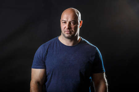 A portrait of pumped up hairless caucasian man in black t-shirt in front of dark background