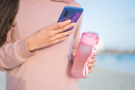 girl holding phone smartphone in hands on the seashore and sport bottle with waterの写真素材