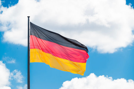 The flag of Germany waving against a bright blue sky with fluffy white clouds, representing German national pride and patriotism.