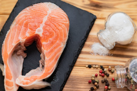 salmon steak on a black slate board on a wooden background view from above next to salt and pepper.の写真素材