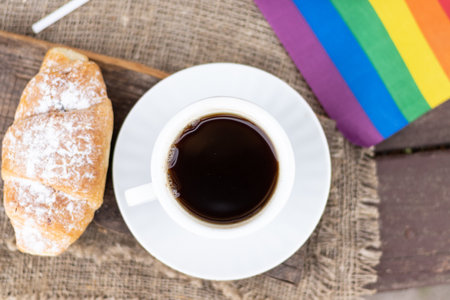 A cup of coffee and a croissant on a wooden table against the background of the LGBT flagの写真素材