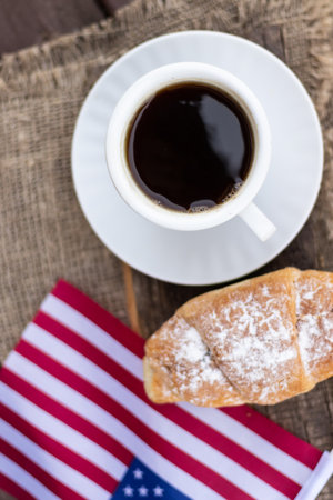 A cup of coffee and a croissant on a wooden table against the background of the American flagの写真素材