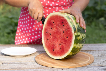 A woman cuts a delicious ripe watermelon at a wooden table on a wooden board outdoors, close-upの写真素材