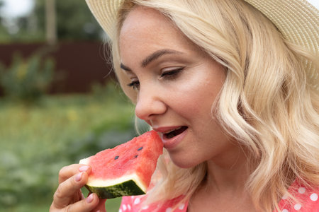 Photo of a young blonde woman in a hat and pink dress. who eats juicy delicious watermelon, biting off pieces of her favorite fruit. Daylight saving time and the concept of nutrition.の写真素材