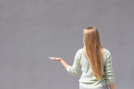 caucasian white girl with long natural blond hair is standing near grey wall and pointing left with...