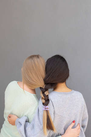 two girls with different color of hair are standing and hugging each other, grey background wall,...